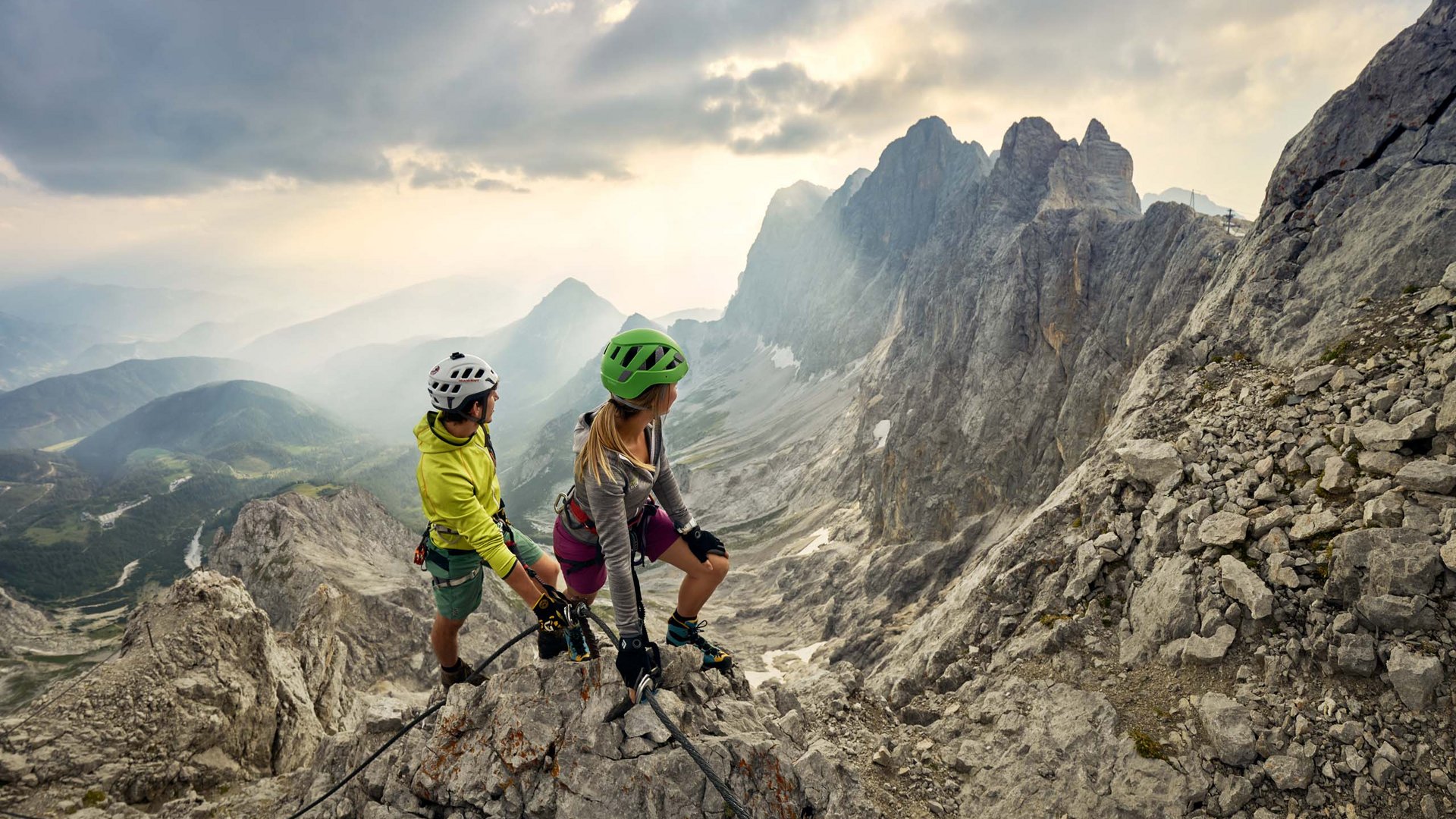 Zwei Kletterer mit Helmen und Kletterausrüstung auf einem Berggipfel mit atemberaubender Aussicht